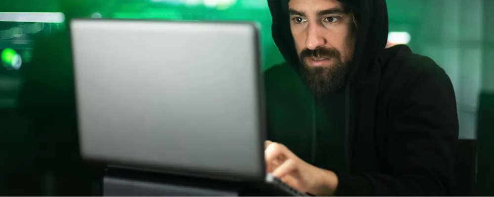 A mysterious person in a hoodie on a computer looking at code to complete a phishing attack.
