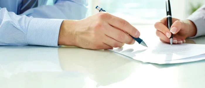 a man wearing a button down and a woman's manicured hand holding a pen and signing merchant agreement documents on a table