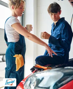 Two auto mechanics in blue coveralls taking a coffee break while chatting near a car with the hood open.