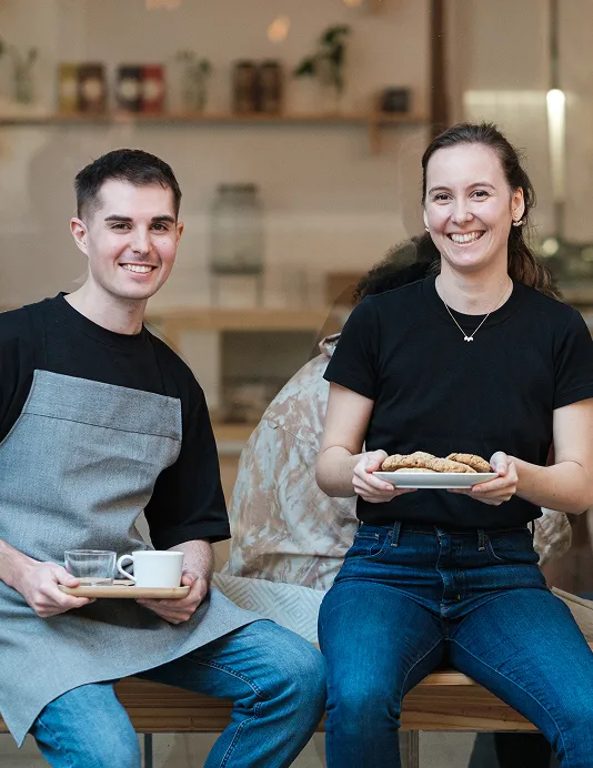 A merchant in an apron and a customer sitting on a table with plates of cookies and beverages, at a coffee shop.