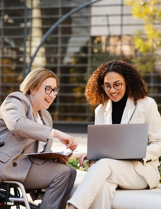 Two smiling businesswomen in pantsuits sitting in front of an office building, one with a notebook and one with a laptop.