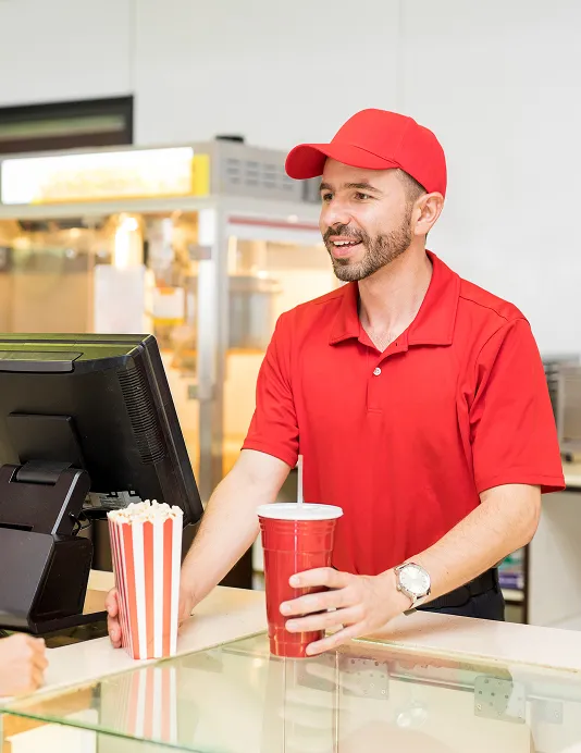 A quick service restaurant worker in a red shirt and had holding a cup and popcorn bag behind a cash register.