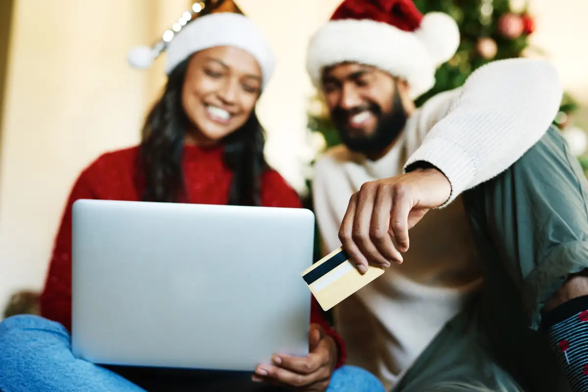 a couple wearing Santa hats and sweaters doing eCommerce shopping on a laptop with a credit card for holiday payment processing