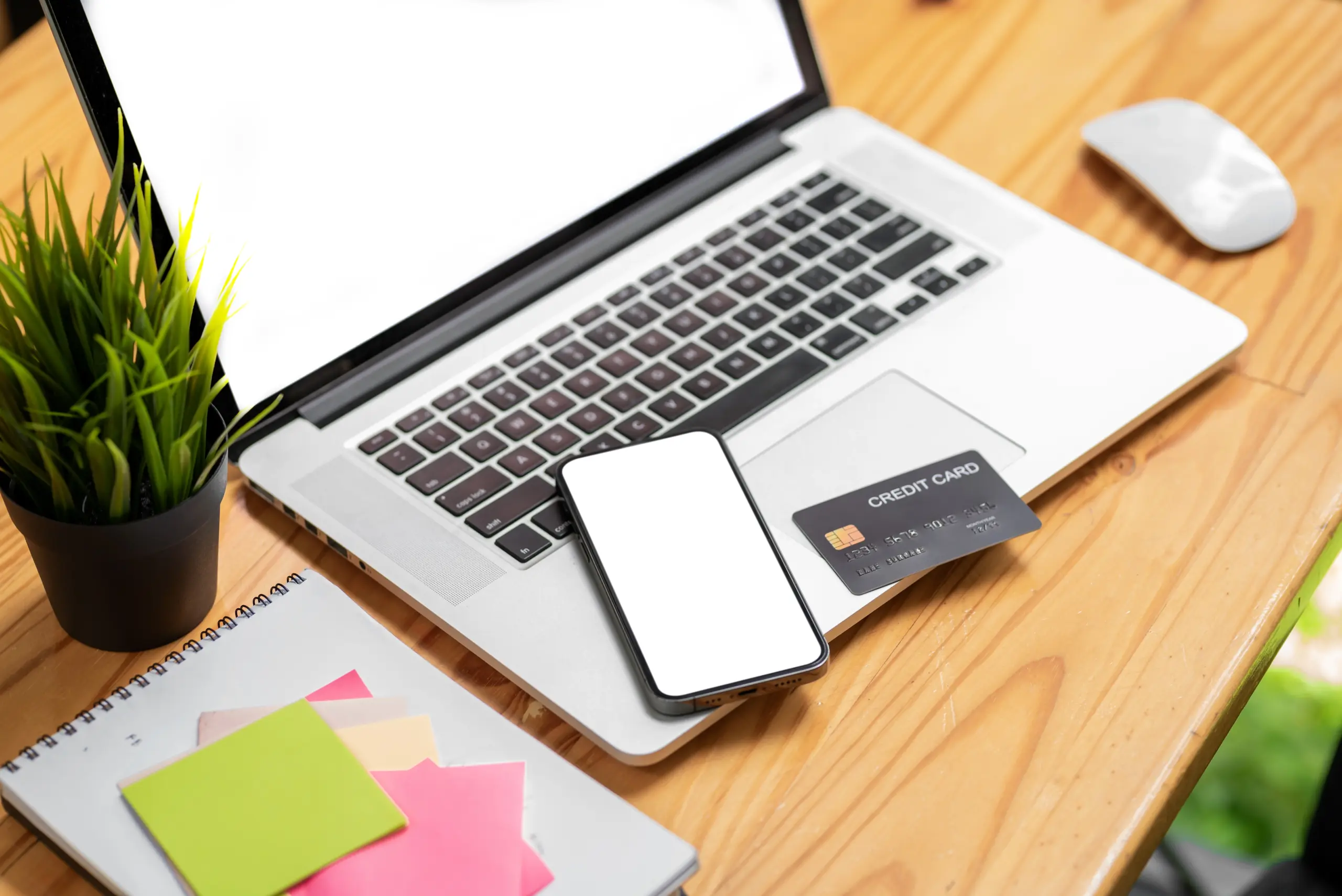 a laptop with mouse and notebook on a desk with a mobile phone and credit card on top of keyboard.